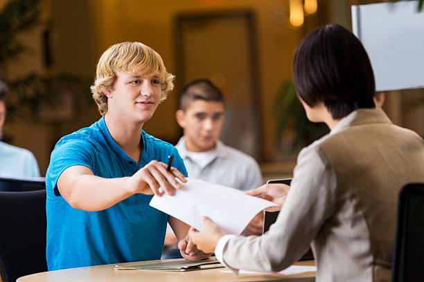 Young college man hands his resume to potential employer at college job fair. Other people are interviewing in the background.