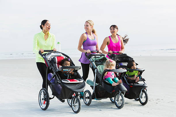 Three multi-ethnic mothers walking together on the beach, pushing their children in jogging strollers.  The child in the middle, a 22 month old toddler with blond hair, is the oldest.  The African American girl and Asian boy are 11 months old.